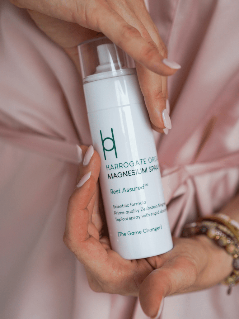 Close-up of hands holding a bottle of Harrogate Organics Magnesium Spray. The person is dressed in a silky soft pink robe, with neatly manicured light pink nails, complementing the product's sleek design.