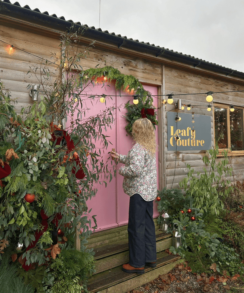 The marketing manager from Harrogate Organics stands outside the Leafy Couture wreath-making workshop. The cosy building features a pink door framed by lush greenery and natural plants. Warm fairy lights drape across the wall and door.